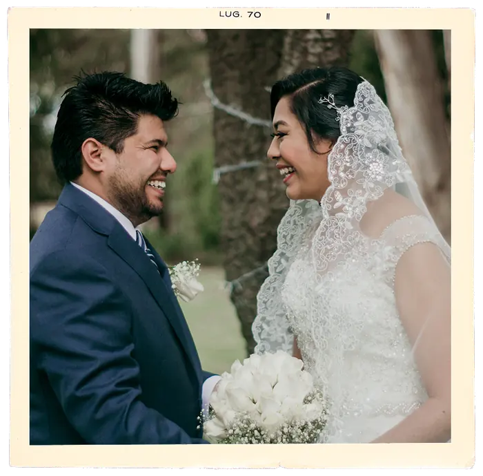 Los novios riendo de frente, él de traje azul, ella con velo de encaje